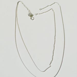 Op de afbeelding: Een dunne, zilverkleurige ketting met een delicaat, uniform schakelontwerp. De ketting is voorzien van een kleine, ronde veerringsluiting aan de bovenkant, waardoor deze veilig kan worden vastgemaakt. Het metaal heeft een heldere afwerking.