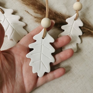 May include: Three white ceramic oak leaf ornaments with a brown suede cord. The leaves have a raised vein pattern and are hanging from a wooden bead.