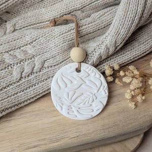 Op de afbeelding: Een witte, ronde keramische luchtverfrisser met een bloemenpatroon, hangend aan een bruin koord en een houten kraal. De luchtverfrisser wordt gepresenteerd tegen een neutrale, getextureerde achtergrond, wat een gezellige, natuurlijke uitstraling creëert.