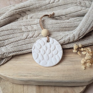 Peut inclure: Un diffuseur d'huiles essentielles rond en céramique blanche avec un motif de galets. Il est suspendu par un cordon en cuir et une perle en bois. Le diffuseur repose sur une surface en bois, avec une écharpe beige en tricot torsadé en arrière-plan.