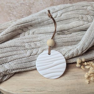 Peut inclure: Un pendentif rond en céramique blanche, texturé avec un motif rayé, suspendu par un cordon en cuir marron et une perle en bois. Il repose sur un tricot torsadé neutre et une surface en bois.