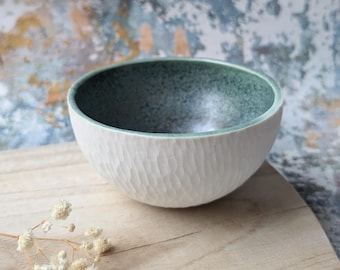 May include: A white ceramic bowl with a textured exterior and a green interior. The bowl is set on a wooden surface, showcasing its handcrafted appearance. The bowl's design is simple and elegant, suitable for serving food or as a decorative piece.