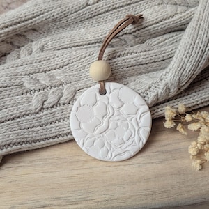 Puede incluir: Un difusor de aromaterapia blanco y circular con un diseño floral en relieve. Cuelga de un cordón de cuero marrón y una cuenta de madera. El difusor se muestra sobre un jersey de punto de cable gris claro.