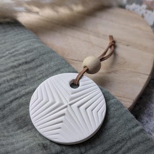 Peut inclure: Un diffuseur d'huiles essentielles rond en céramique blanche avec un motif géométrique en forme d'étoile. Il est suspendu par un cordon en cuir marron et une perle en bois. Le diffuseur repose sur un tissu vert et une surface en bois.