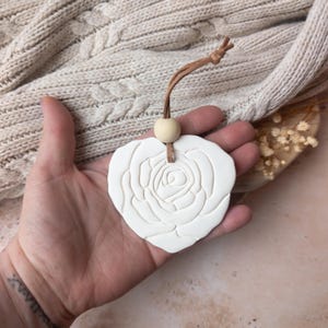 Könnte beinhalten: Ein weißer, herzförmiger Lufterfrischer mit Rosenmuster. Er hat eine braune Lederschnur und eine Holzkugel zum Aufhängen. Der Lufterfrischer wird in einer Hand vor einem neutralen Hintergrund gehalten.