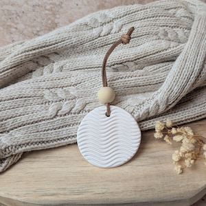 Op de afbeelding: Een witte, cirkelvormige aromatherapie-diffuser met een golfpatroon, opgehangen aan een bruin leren koord en een houten kraal. De diffuser rust op een houten oppervlak, met een neutraal gekleurde gebreide deken op de achtergrond.
