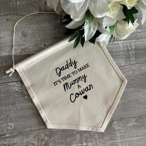 Puede incluir: Una pancarta de lona blanca con texto negro que dice "Daddy It's Time To Make Mummy A Cowan" con un pequeño corazón negro.