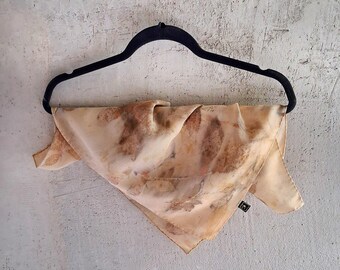 May include: A beige square scarf with brown and orange leaf patterns hangs from a black hanger against a gray wall. The scarf has a natural, organic design, and a small tag is visible.