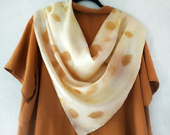 May include: A cream-colored silk scarf with a leaf pattern in shades of brown and tan. The scarf is draped over a rust-colored dress, creating a layered effect. The scarf is square-shaped and appears to be handmade.