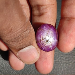 May include: A purple star ruby gemstone with a star-shaped pattern in the center. The gemstone is oval-shaped and has a smooth, polished surface.