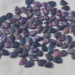 May include: A collection of approximately 50 purple and black teardrop-shaped gemstone cabochons. The stones are smooth and polished, with a variety of patterns and swirls.