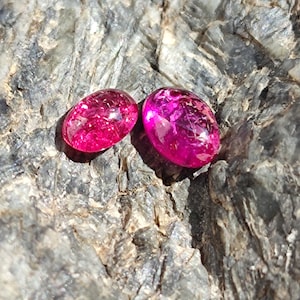 Op de afbeelding: Twee ovaalvormige, levendige roze edelstenen met interne insluitsels worden getoond op een getextureerd, grijs rots oppervlak. De edelstenen hebben een gepolijste, glanzende uitstraling, die licht reflecteert en hun rijke kleur laat zien.