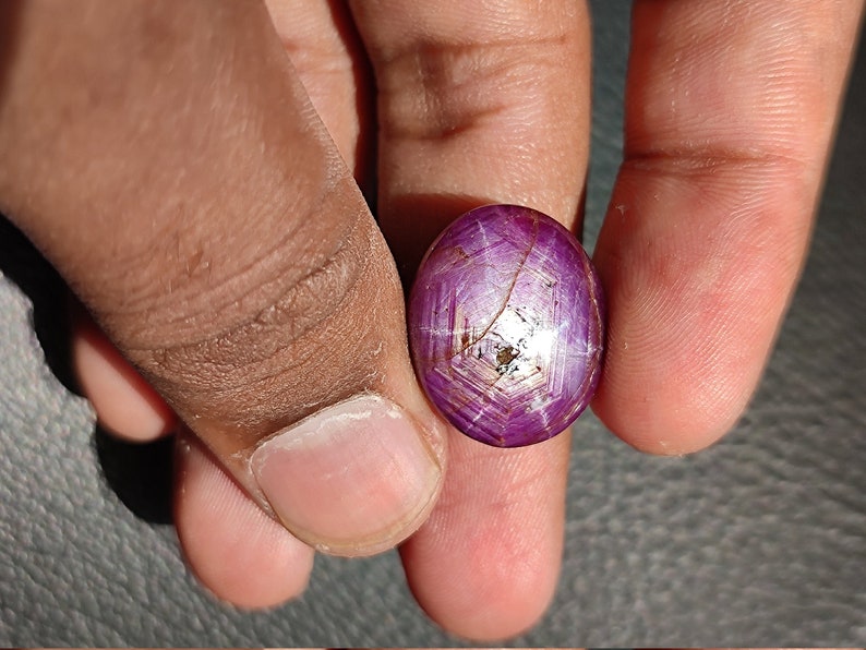 May include: A purple star ruby gemstone with a star-shaped pattern in the center. The gemstone is oval-shaped and has a smooth, polished surface.