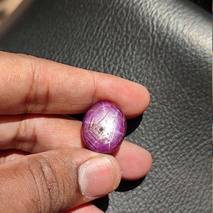 May include: A close-up of a pink oval-shaped gemstone with a star-like pattern and a silver-colored inclusion. The gemstone is held in a hand.