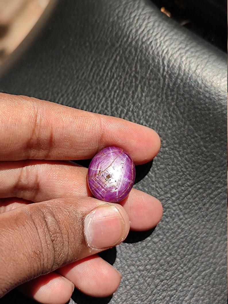May include: A close-up of a pink oval-shaped gemstone with a star-like pattern and a silver-colored inclusion. The gemstone is held in a hand.