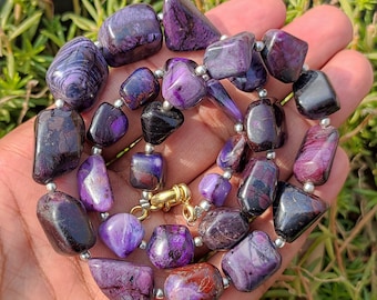 Puede incluir: Un collar de piedras preciosas de color púrpura y negro con cuentas de plata. El collar está hecho de piedras de formas irregulares y tiene aproximadamente 45 cm de largo.