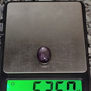 May include: A dark purple gemstone with a star-like pattern is displayed on a digital scale. The scale reads 5.360 ct.