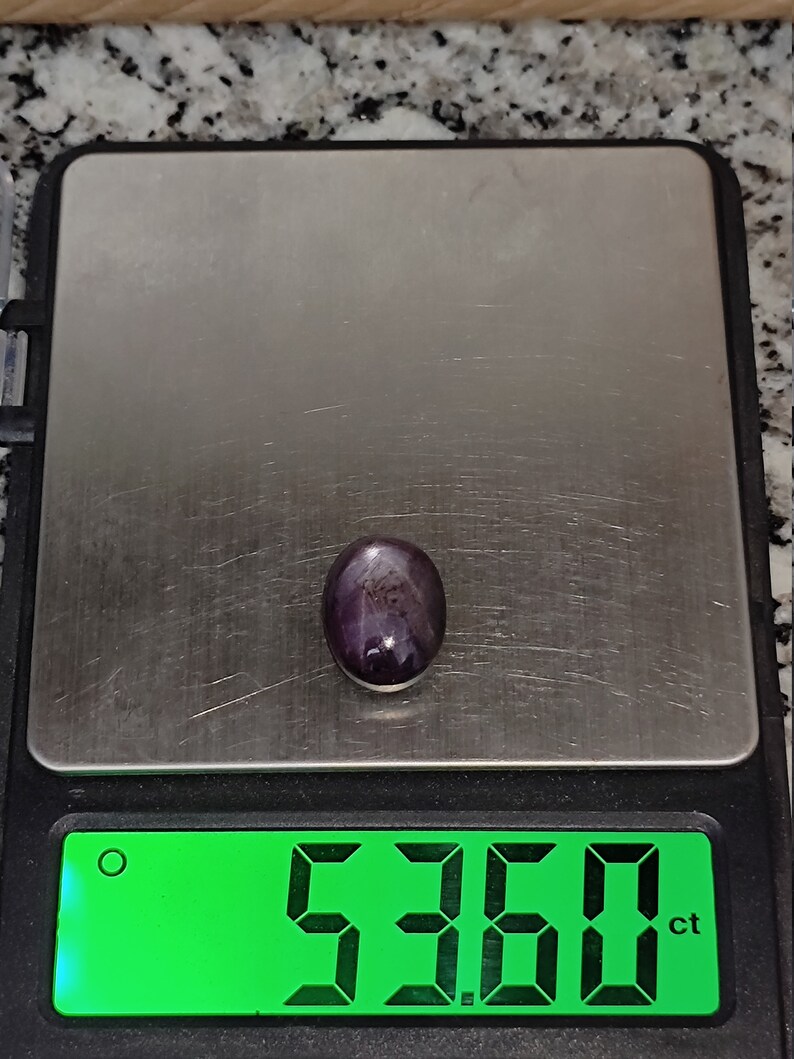 May include: A dark purple gemstone with a star-like pattern is displayed on a digital scale. The scale reads 5.360 ct.