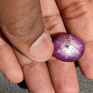 May include: A close-up of a round, purple gemstone with a star-shaped pattern. The gemstone is held in a hand.