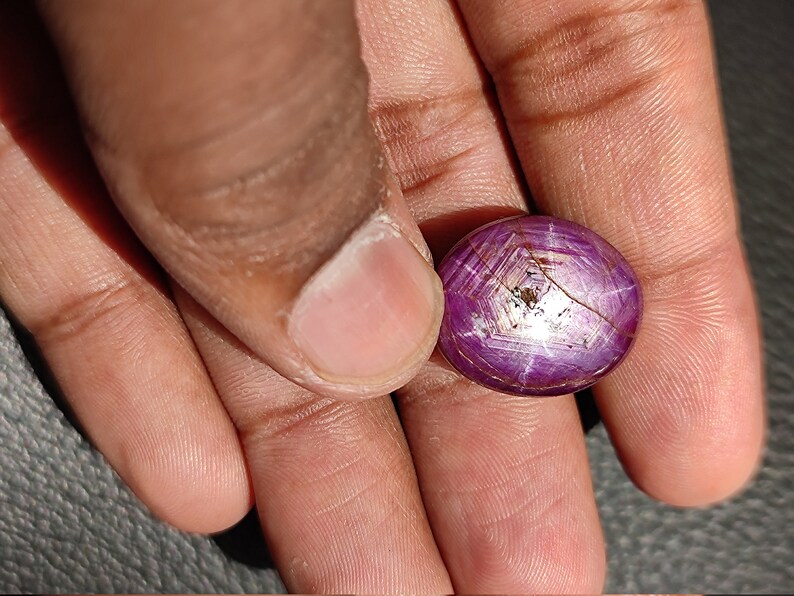 May include: A close-up of a round, purple gemstone with a star-shaped pattern. The gemstone is held in a hand.