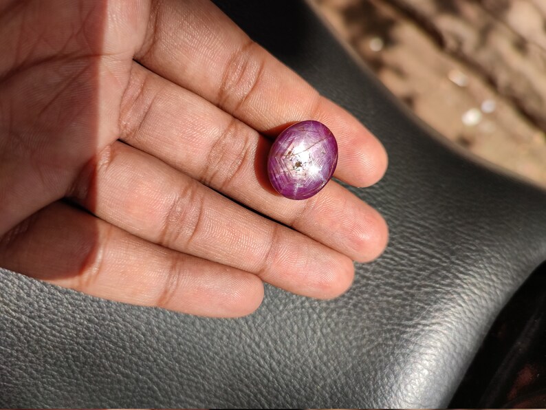May include: A pink oval-shaped gemstone with a star-like pattern inside. The gemstone is set in a silver ring.