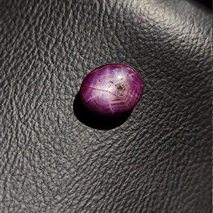May include: A deep red oval-shaped gemstone with a star-like pattern inside. The gemstone is set against a dark, textured background.
