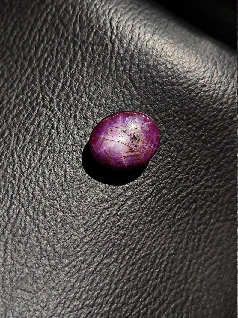 May include: A deep red oval-shaped gemstone with a star-like pattern inside. The gemstone is set against a dark, textured background.