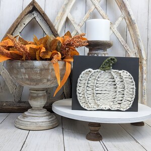 May include: A black rectangular wooden sign featuring a string art pumpkin design. The pumpkin is created with thick cream-colored yarn wrapped around small silver nails, with a green yarn stem. The sign is displayed on a white wooden pedestal stand.