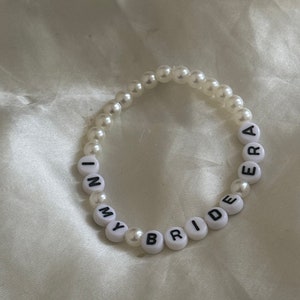 May include: A white pearl bracelet with the words "In My Bride Era" spelled out in black letters.
