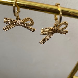 May include: A pair of gold hoop earrings with a delicate bow charm. The bows are made of intricate, woven metal and have a slightly textured finish.