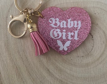 May include: Pink glitter heart-shaped keychain with a gold key ring and a pink tassel. The heart has a white butterfly and the text "Baby Girl" in white.