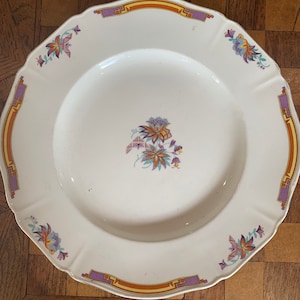 May include: A white ceramic plate with a decorative floral design in shades of purple, orange, and yellow. The plate has a scalloped edge and a raised rim.