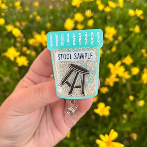 May include: A teal blue glitter badge holder with a white label that says "Stool Sample". Inside the holder is a black silhouette of a stool.