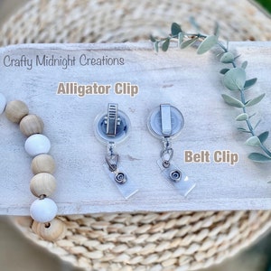 May include: Two silver retractable badge holders with clear plastic cases. The holder on the left has an alligator clip attachment and the holder on the right has a belt clip attachment. The text "Alligator Clip" is above the left holder and the text "Belt Clip" is above the right holder.