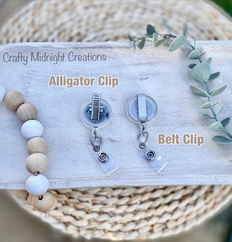 May include: Two silver retractable badge holders with clear plastic cases. The holder on the left has an alligator clip attachment and the holder on the right has a belt clip attachment. The text "Alligator Clip" is above the left holder and the text "Belt Clip" is above the right holder.