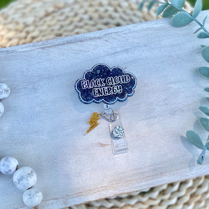 May include: A black glitter cloud-shaped badge holder with the text "Black Cloud Energy" in white lettering. A gold lightning bolt charm is attached to the badge holder.