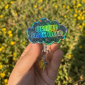 May include: A black glitter cloud-shaped badge holder with a silver retractable badge reel and a silver lightning bolt charm. The badge holder says "CERTIFIED BLACK CLOUD".