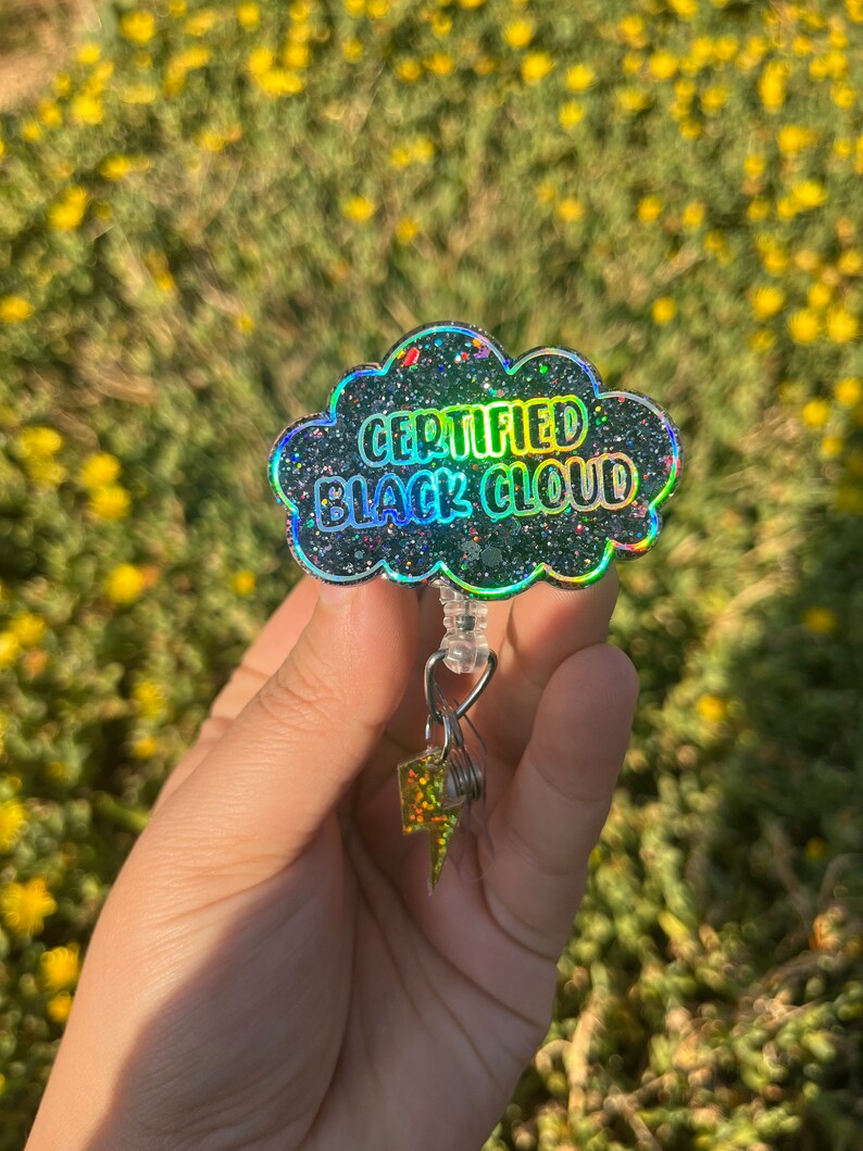 May include: A black glitter cloud-shaped badge holder with a silver retractable badge reel and a silver lightning bolt charm. The badge holder says "CERTIFIED BLACK CLOUD".