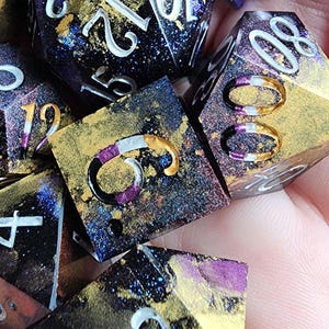 May include: A set of six polyhedral dice with a galaxy-inspired design. The dice are black with gold, purple, and white accents. The numbers on the dice are white and outlined in silver. The dice are made of resin and have a glittery finish.