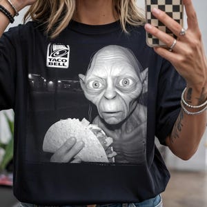 Puede incluir: Camiseta negra con una imagen en escala de grises de una criatura sosteniendo un taco, y el logo de Taco Bell. El diseño tiene una cualidad humorística y surrealista, mezclando la cultura pop con elementos fantásticos.