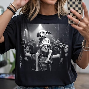 Puede incluir: Camiseta negra con una imagen en escala de grises de una multitud en un concierto o fiesta. La imagen presenta a una persona sobre los hombros, con gafas y un gorro, y una criatura con orejas grandes.