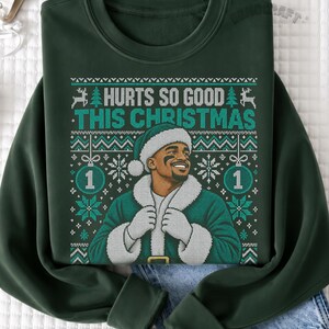 Op de afbeelding: Een donkergroene sweater met ronde hals en een grafische print van een persoon in een kerstmanpak met de tekst "HURTS SO GOOD THIS CHRISTMAS" en het cijfer "1" in twee ornamenten, omringd door een blauwgroene en witte kerstrand.