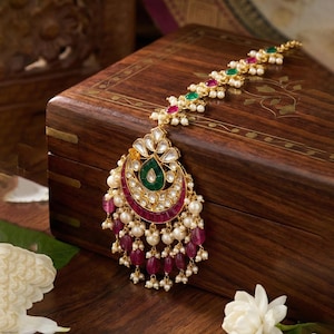 May include: An ornate gold-toned headpiece with a central crescent design, featuring a green teardrop-shaped stone, surrounded by red and white stones. The headpiece is adorned with pearls and ruby-colored beads, and green and red accents.