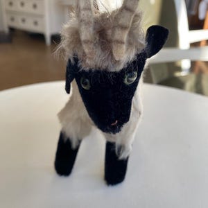 Könnte beinhalten: Ein Vintage-Ziegen-Stofftier mit schwarzem Gesicht und Beinen, beigefarbenem Fell und kleinen, gebogenen Hörnern. Die Ziege hat grüne Augen und steht auf einer weißen Oberfläche. Das Spielzeug ist ein Sammlerstück.