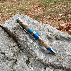 Pode incluir: Uma caneta artesanal com um corpo de madeira com uma mistura de tons castanhos claros e escuros, acentuada com faixas azuis e douradas. A caneta tem um clipe e uma ponta prateados, repousando sobre uma rocha cinzenta.