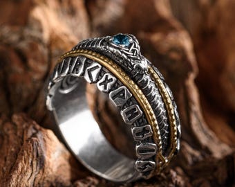 May include: A silver ring with intricate detailing, including a snake design and a small blue gemstone. The ring has gold-coloured accents and engraved lettering. The ring is presented on a wooden surface.