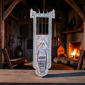 Puede incluir: Una lira de madera con diseño vikingo. El instrumento presenta una caja de resonancia rectangular con un patrón geométrico tallado y un borde trenzado. La parte superior tiene tallas de cabeza de dragón. El fondo muestra un interior rústico con chimenea.