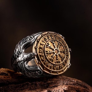 May include: A silver ring with a bronze-coloured central emblem. The ring features a detailed design of a compass-like symbol, surrounded by intricate patterns. The ring's band is adorned with stylized bird or raven figures, adding to its Viking-inspired aesthetic.