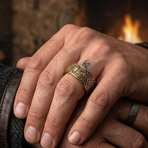 May include: A gold-colored ring with an intricate Celtic knot design. The ring is worn on a finger, showcasing the detailed pattern and texture. A second ring with a braided design is also visible.