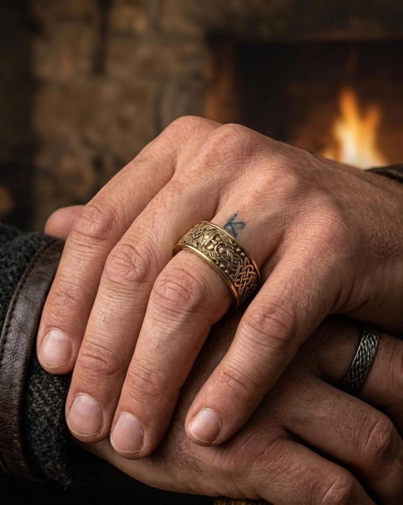 May include: A gold-colored ring with an intricate Celtic knot design. The ring is worn on a finger, showcasing the detailed pattern and texture. A second ring with a braided design is also visible.
